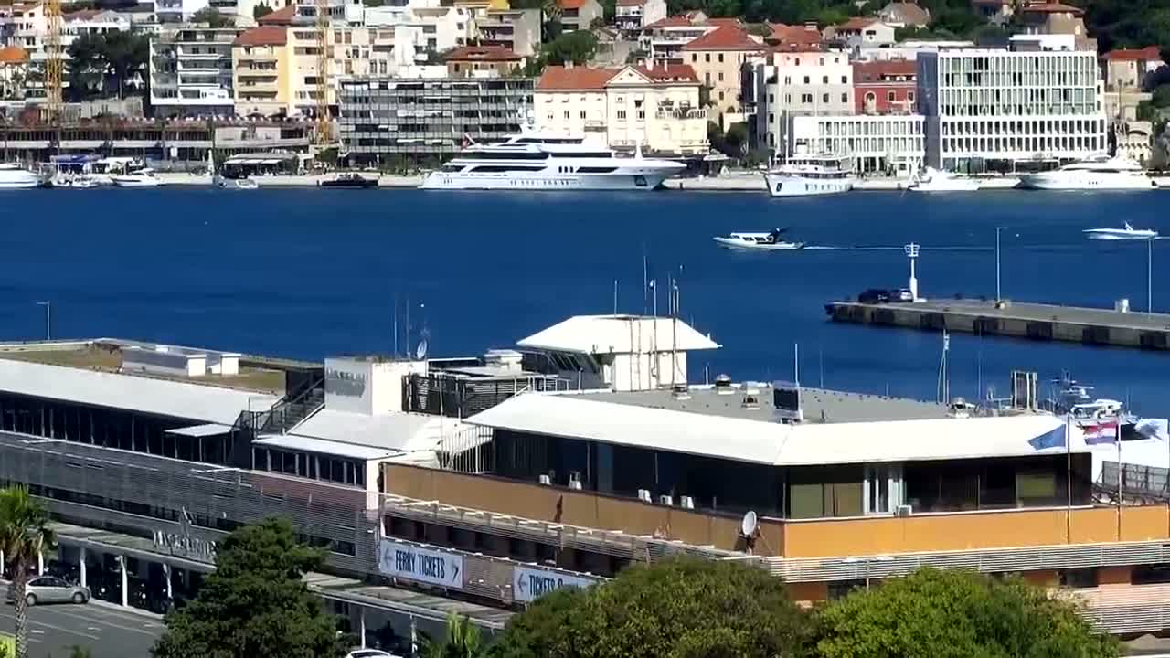 A large building sits on the waterfront overlooking a harbor with numerous boats and yachts, under a clear sunny sky.