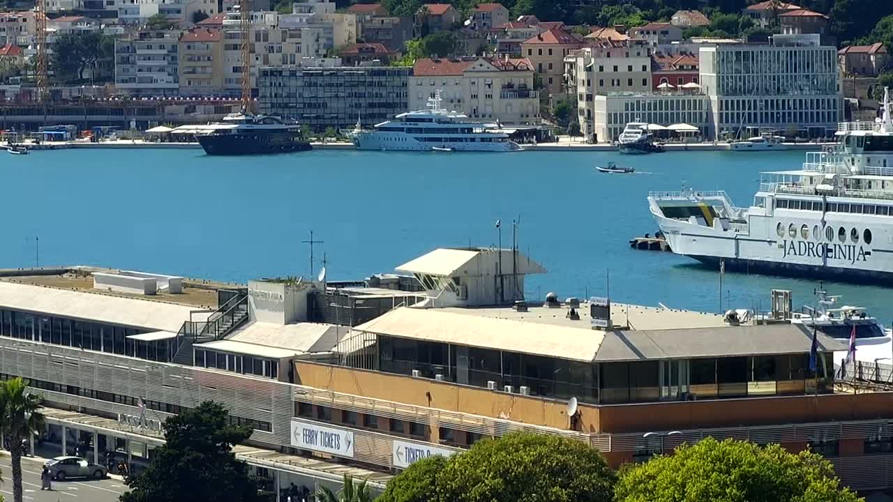 A harbor with several boats and ships, including a large ferry, is seen from an elevated vantage point on a sunny day.