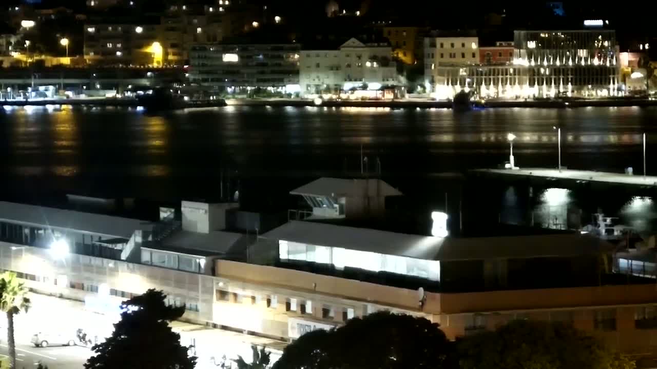 A nighttime view of a city harbor with calm water reflecting the lights of buildings and a dock.