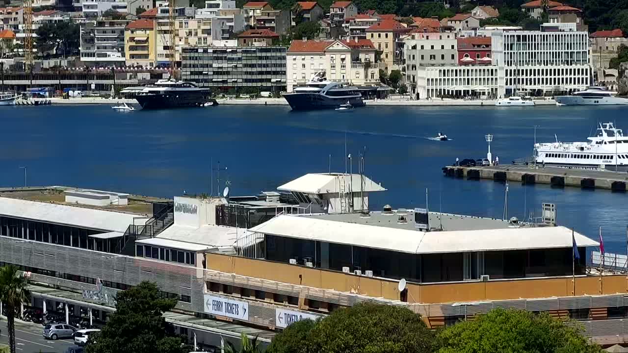 A harbor scene on a sunny day shows a large building near the water's edge, several yachts and a ferry docked, and a city rising up behind the harbor.