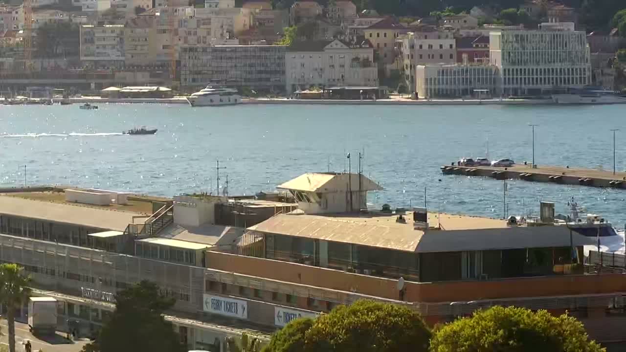 A waterfront building with a flat roof and a control tower overlooks a calm harbor with several boats and a city in the background on a sunny day.