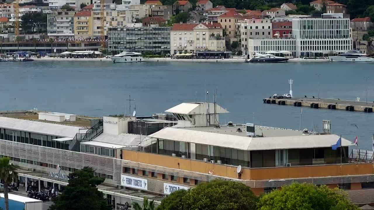 A ferry terminal building with a flat roof sits on the waterfront, overlooking a calm body of water with several boats and a city of multi-story buildings in the background.