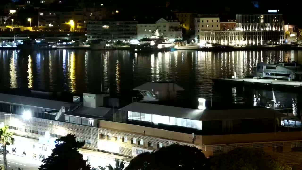 A nighttime view of a calm harbor reflecting the city lights, with boats moored at a dock and buildings along the waterfront.