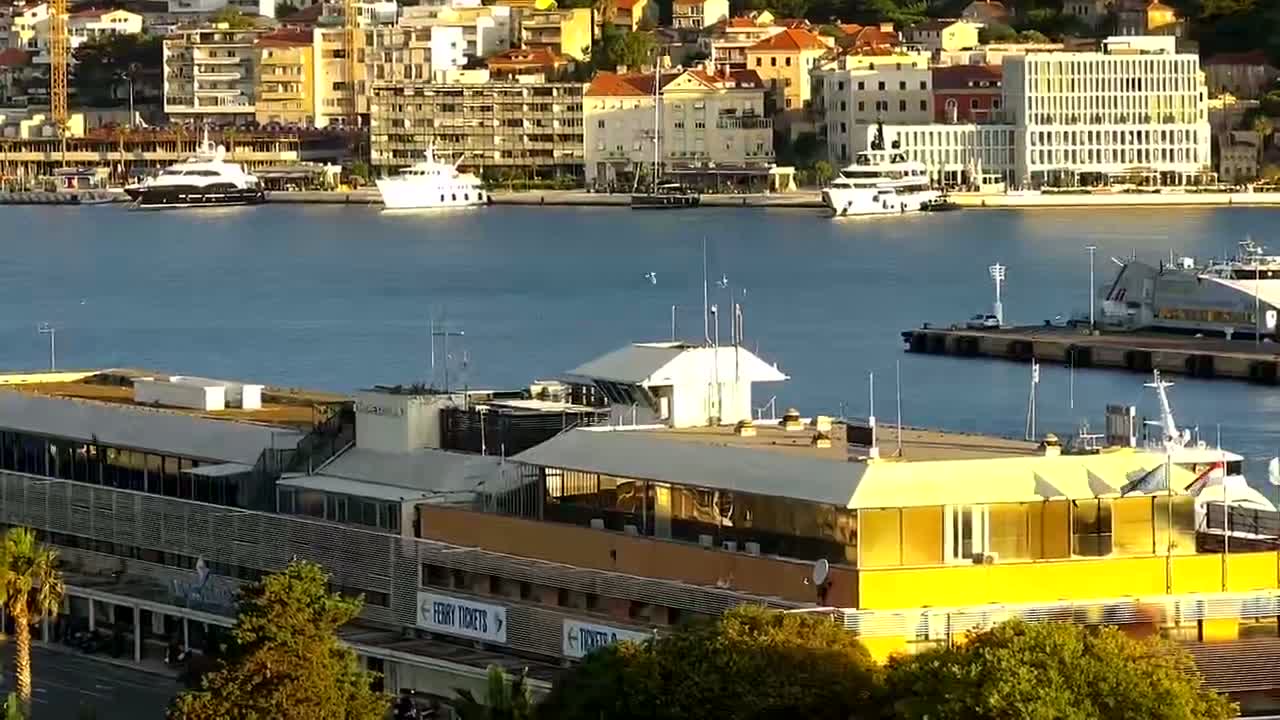 A yellow-roofed building near a calm harbor with several boats and a cityscape in the background under a sunny sky.