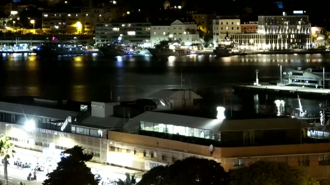 A nighttime view of a harbor with several boats docked, illuminated buildings along the shoreline, and a large building with a flat roof in the foreground.