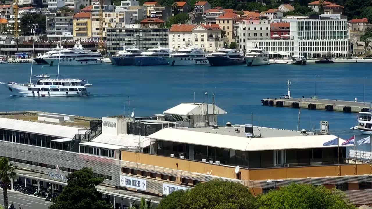 A harbor filled with numerous yachts and boats is seen from above on a sunny day, with a ferry terminal and a city in the background.