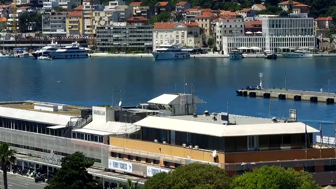 A harbor scene shows a waterfront building with a flat roof, several yachts and boats in the water, and a town of light-colored buildings rising on a hillside in the background under a sunny sky.