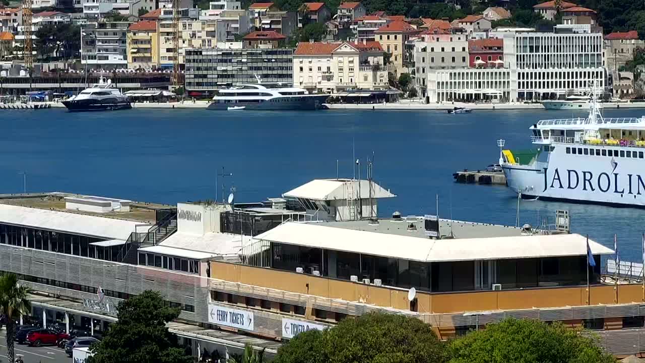 A ferry is docked near a harbor building next to a calm body of water with several yachts and a city in the background on a sunny day.