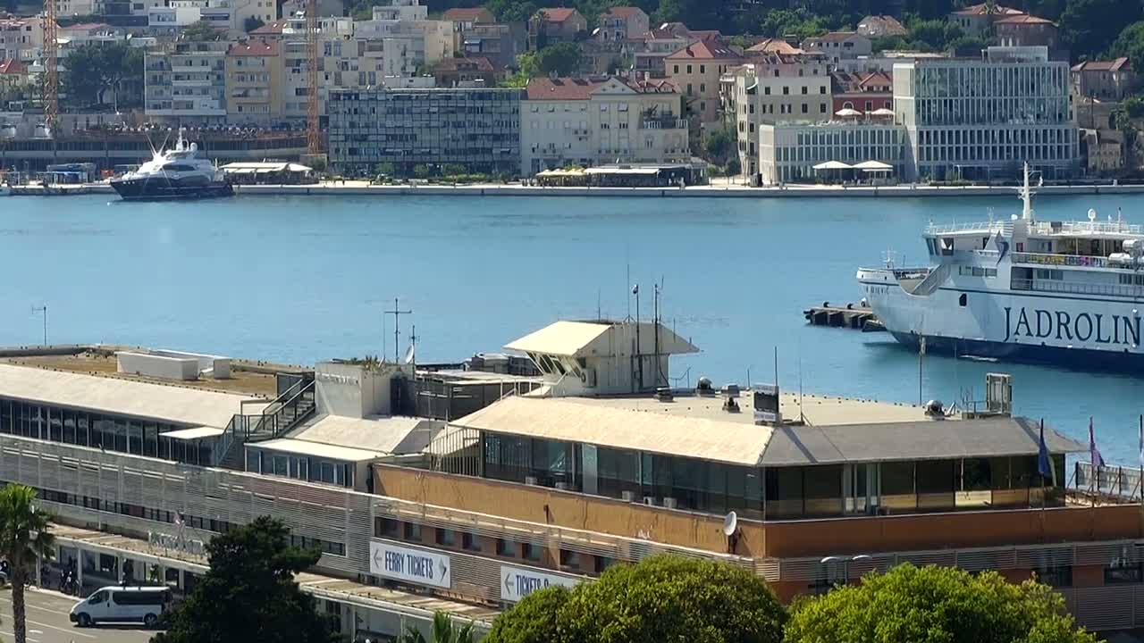 A ferry and a yacht are docked in a harbor next to a large building, with a city and hills visible in the background under a sunny sky.