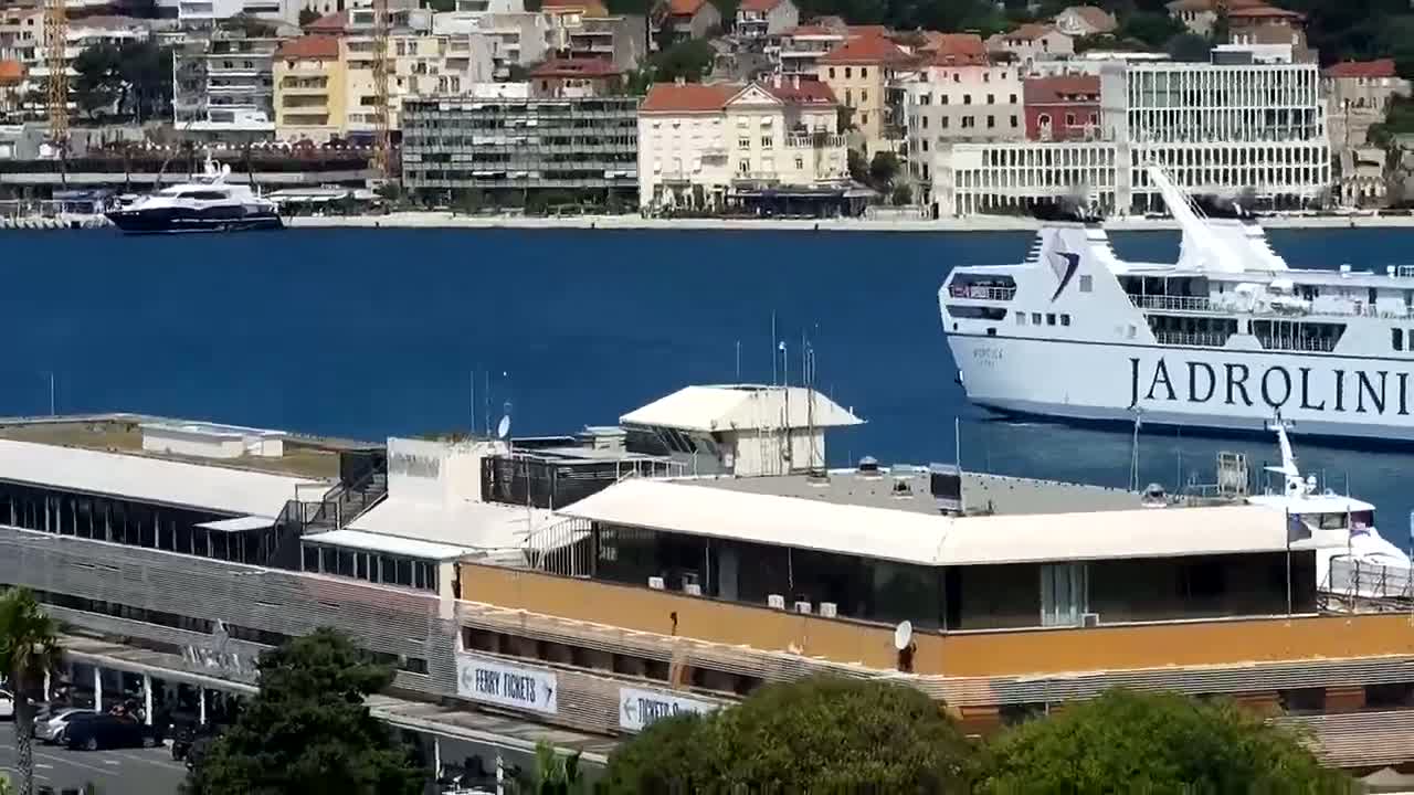 A ferry and a yacht are in a calm blue harbor next to a port building and a city.