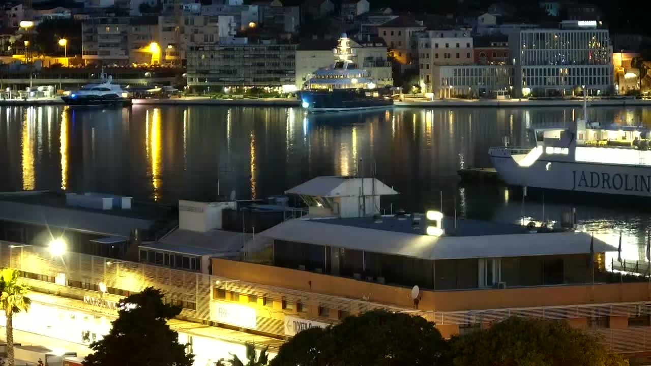 A nighttime harbor scene shows a city's waterfront illuminated, with yachts and ferries moored, reflecting in calm water.