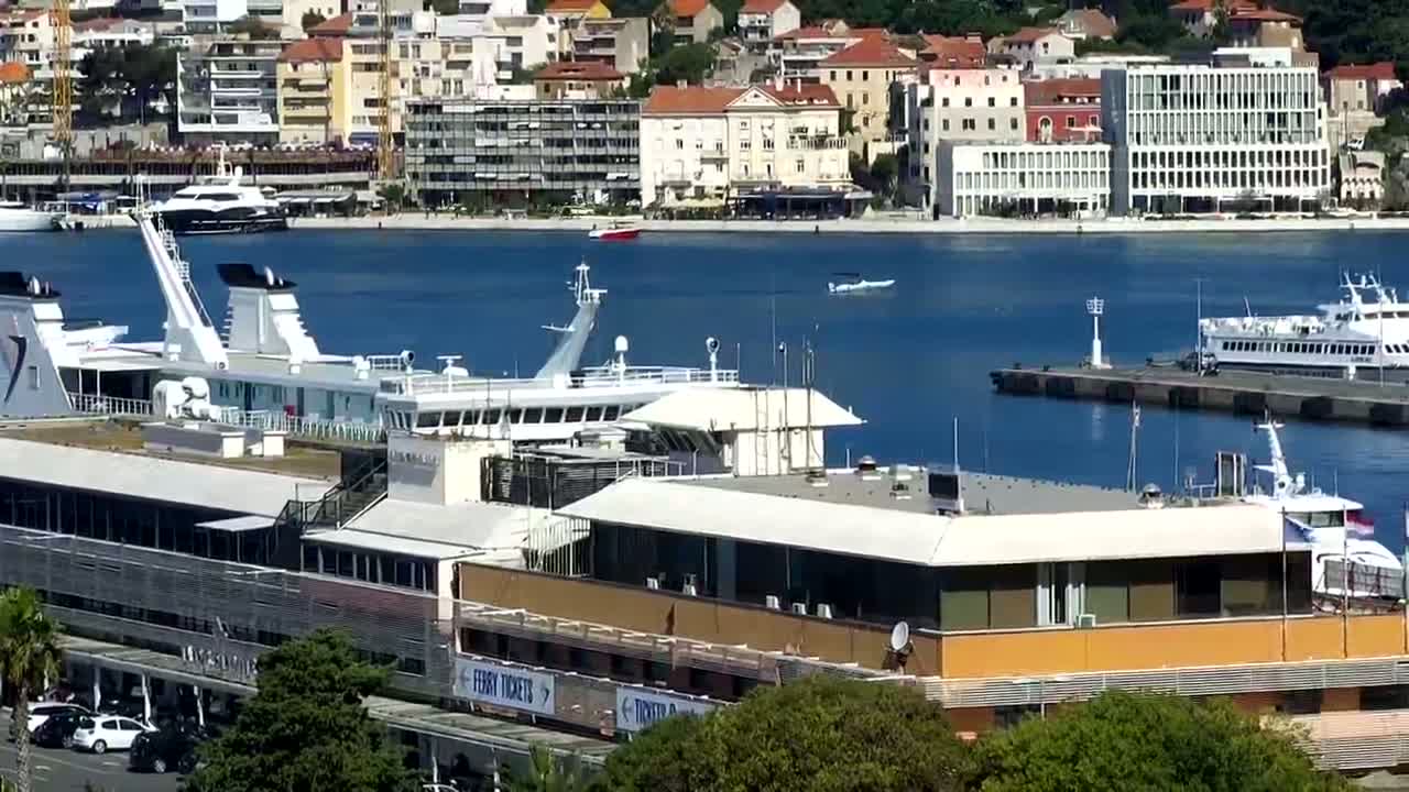 A harbor with several docked ships and boats, a waterfront building, and a city skyline in the background on a sunny day.