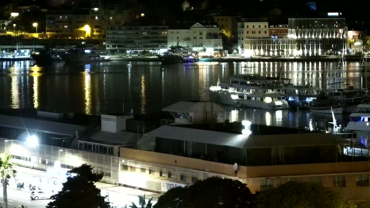 A nighttime view of a harbor filled with boats and yachts, reflecting the lights of buildings along the waterfront under calm conditions.