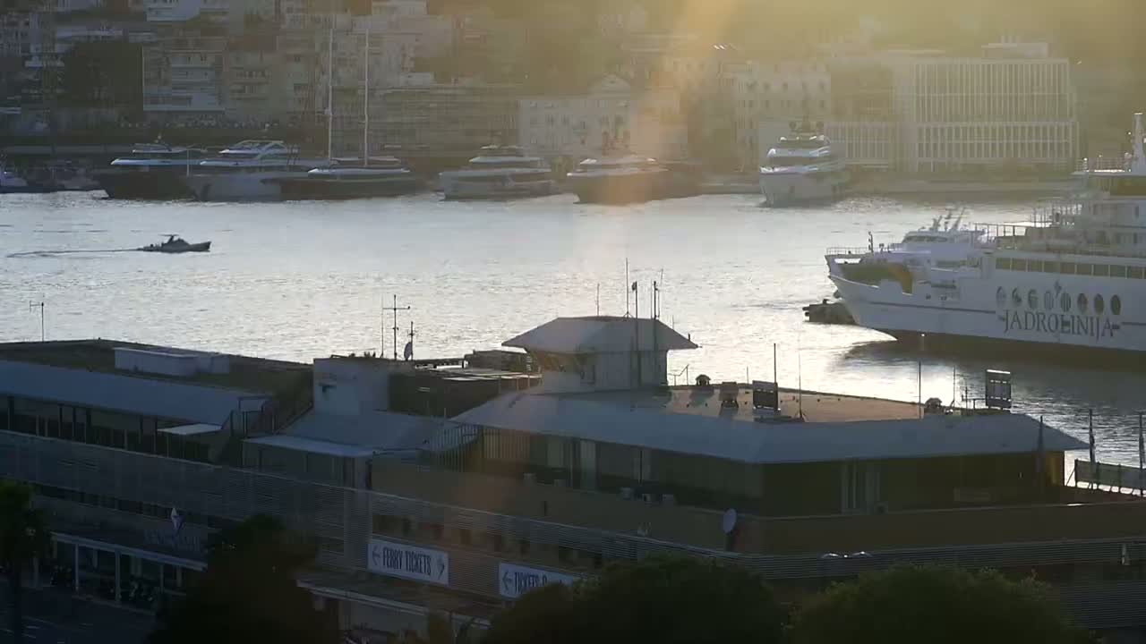 A harbor scene at sunset, showing a ferry, several boats, and a building, with calm water reflecting the golden light.