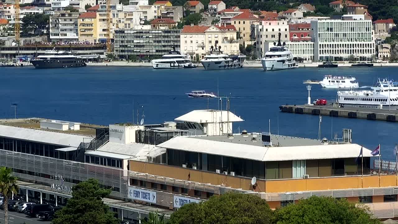 A harbor with several yachts and ferries, a control tower, and a building is seen on a sunny day.