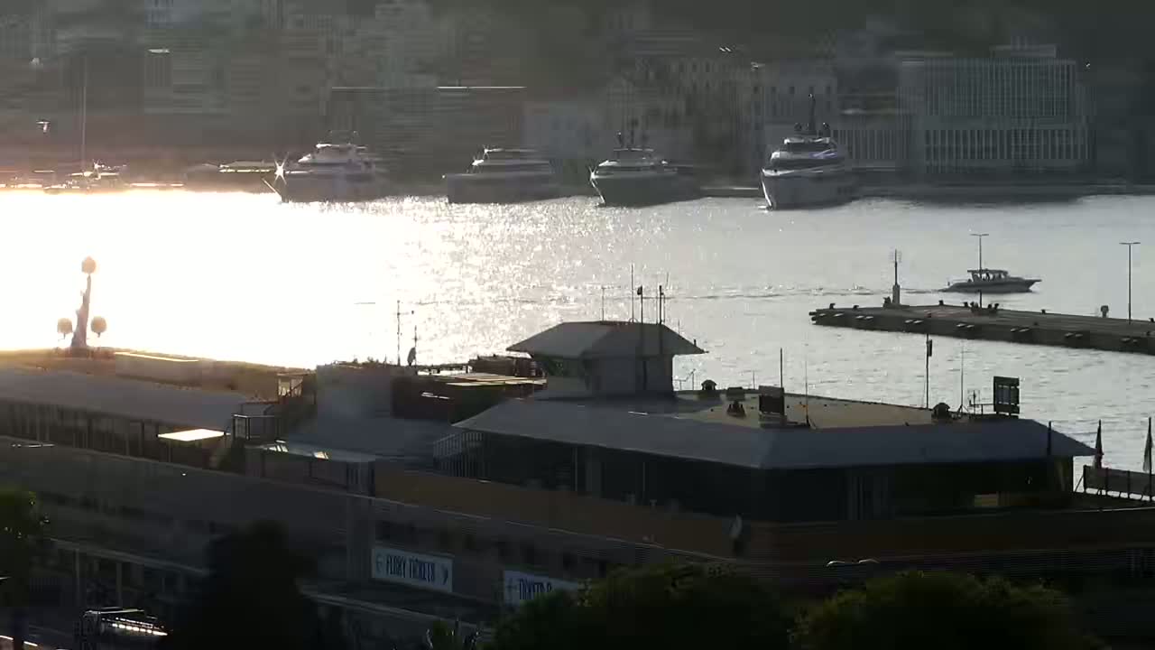 Several boats are moored in a harbor next to a building, with the sun reflecting brightly off the water.