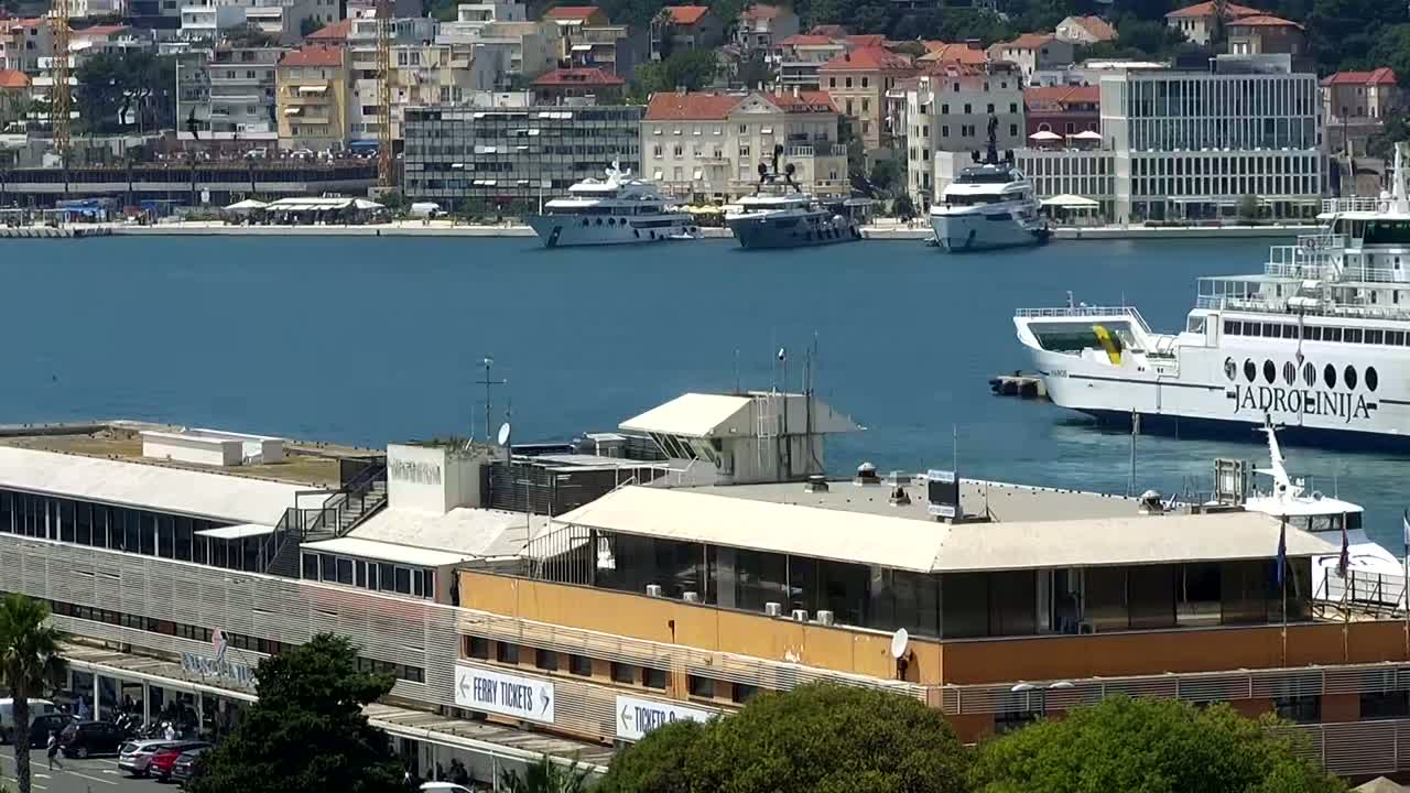 A ferry and several yachts are moored in a harbor next to a multi-story building, under a sunny sky.