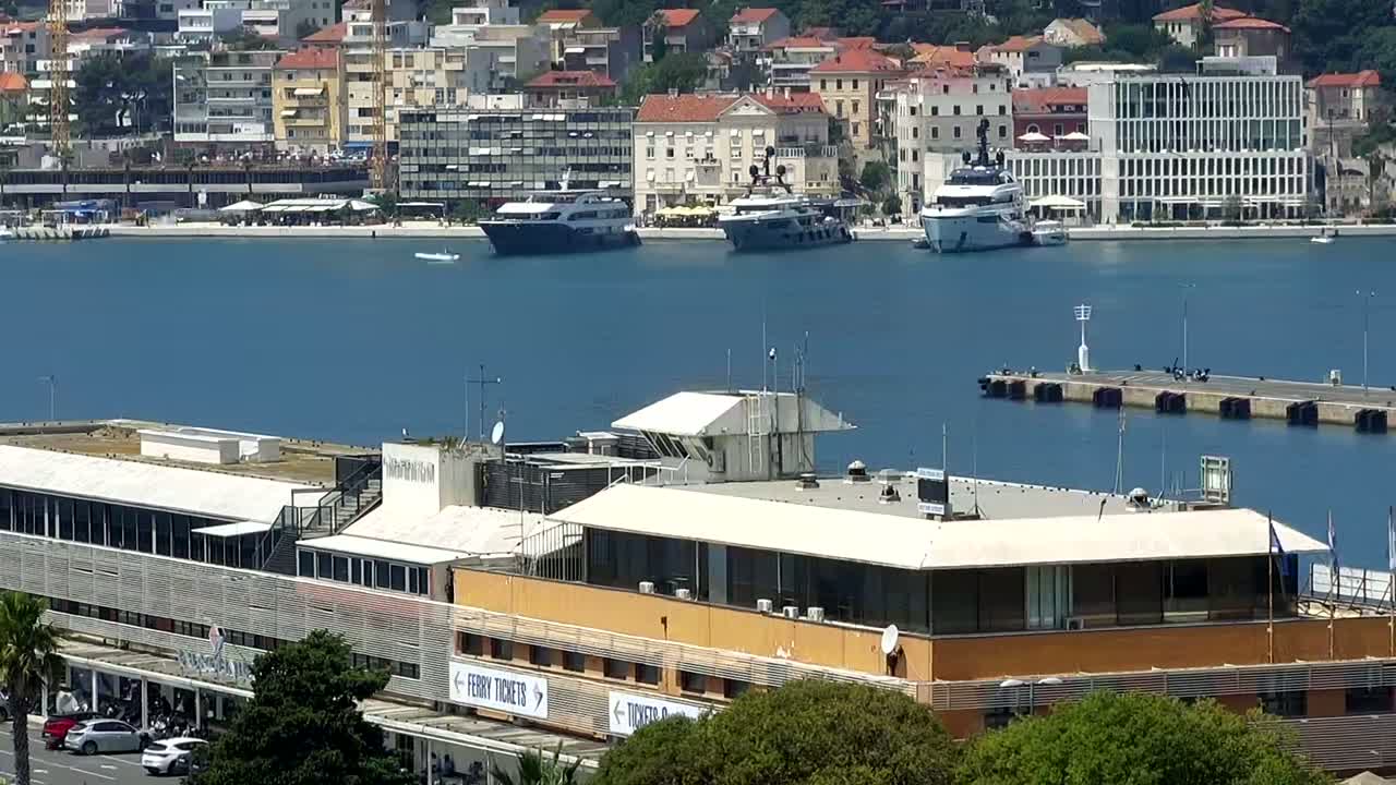 A large building sits by a calm blue body of water, where several yachts are docked near a waterfront city under a clear sky.