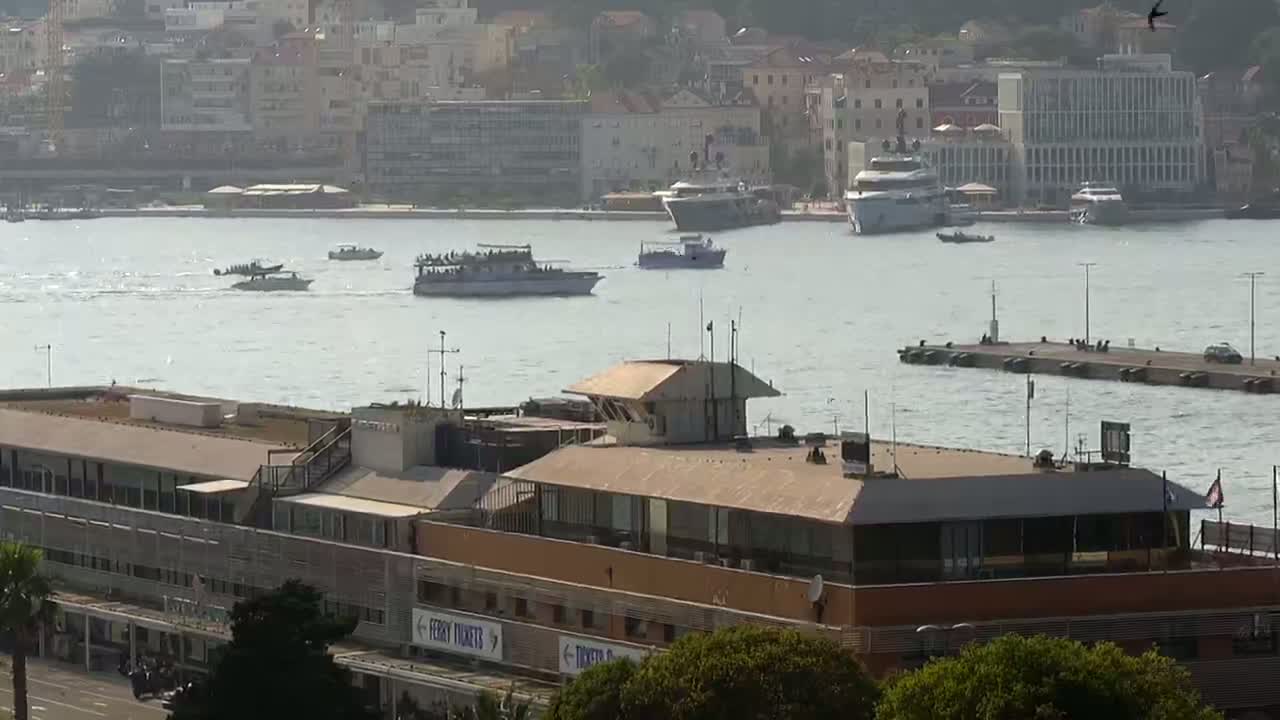 A harbor scene on a partly sunny day shows several boats, a large building with a rooftop control tower, and a city in the background.