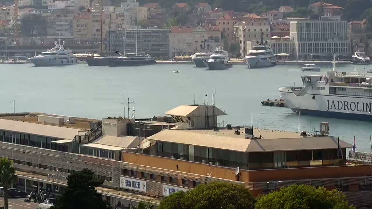 A large ferry and several yachts are moored in a harbor next to a multi-story building, under a mostly clear sky.