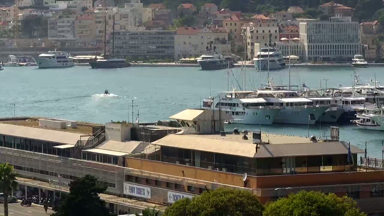 A calm harbor filled with numerous boats and yachts is seen from an elevated vantage point, with a tan-colored building in the foreground.
