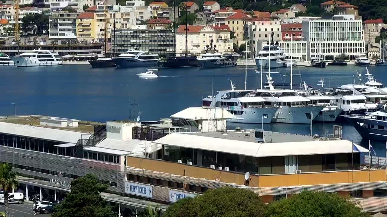 A harbor filled with numerous yachts and boats is viewed from above on a sunny day, with a building in the foreground.