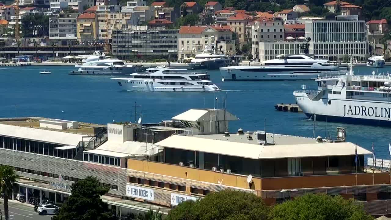 Several yachts and a ferry are moored in a harbor next to a multi-story building on a sunny day.
