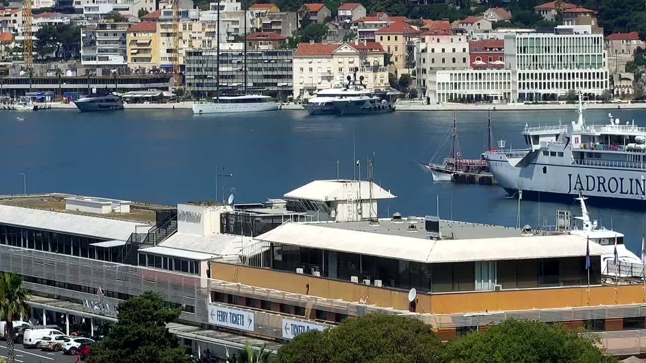 A harbor scene on a sunny day shows several yachts and ferries moored near a building with a flat roof.