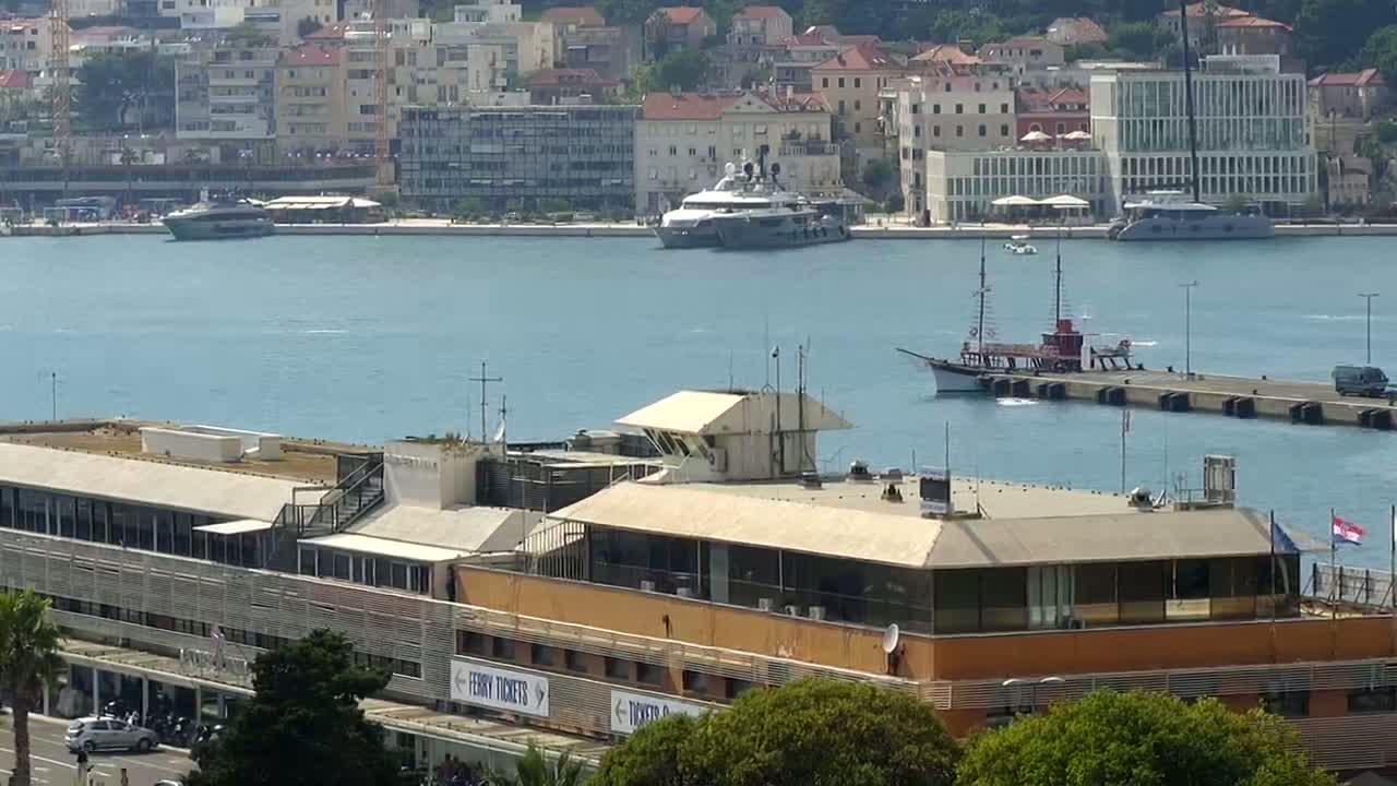 A large building sits on a waterfront, with several boats and yachts in a calm harbor next to a city.