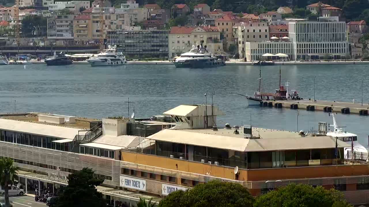 A harbor scene on a sunny day shows several yachts and a ferry docked near a multi-story building.
