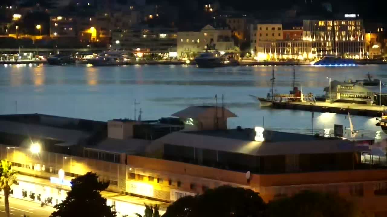 A nighttime view of a harbor with several boats and buildings illuminated along the waterfront under a calm, dark sky.