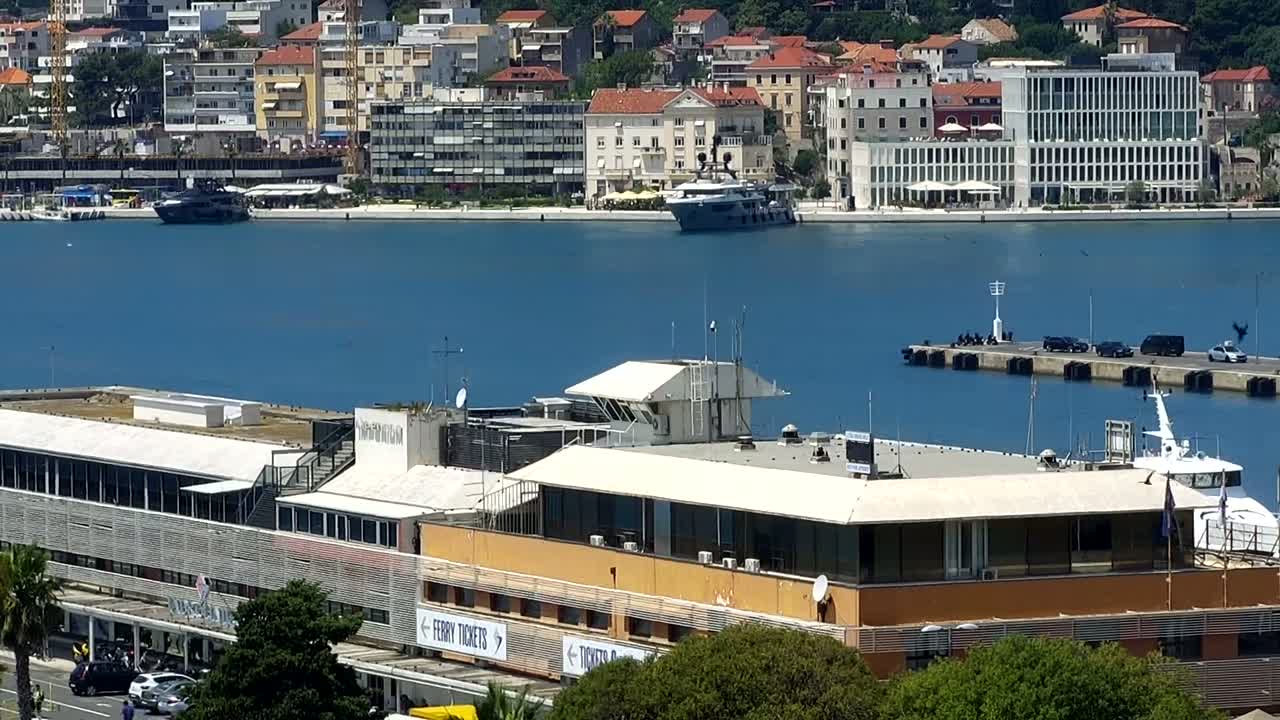 A large building complex near a harbor with a yacht and several cars parked on a dock, under a sunny sky.