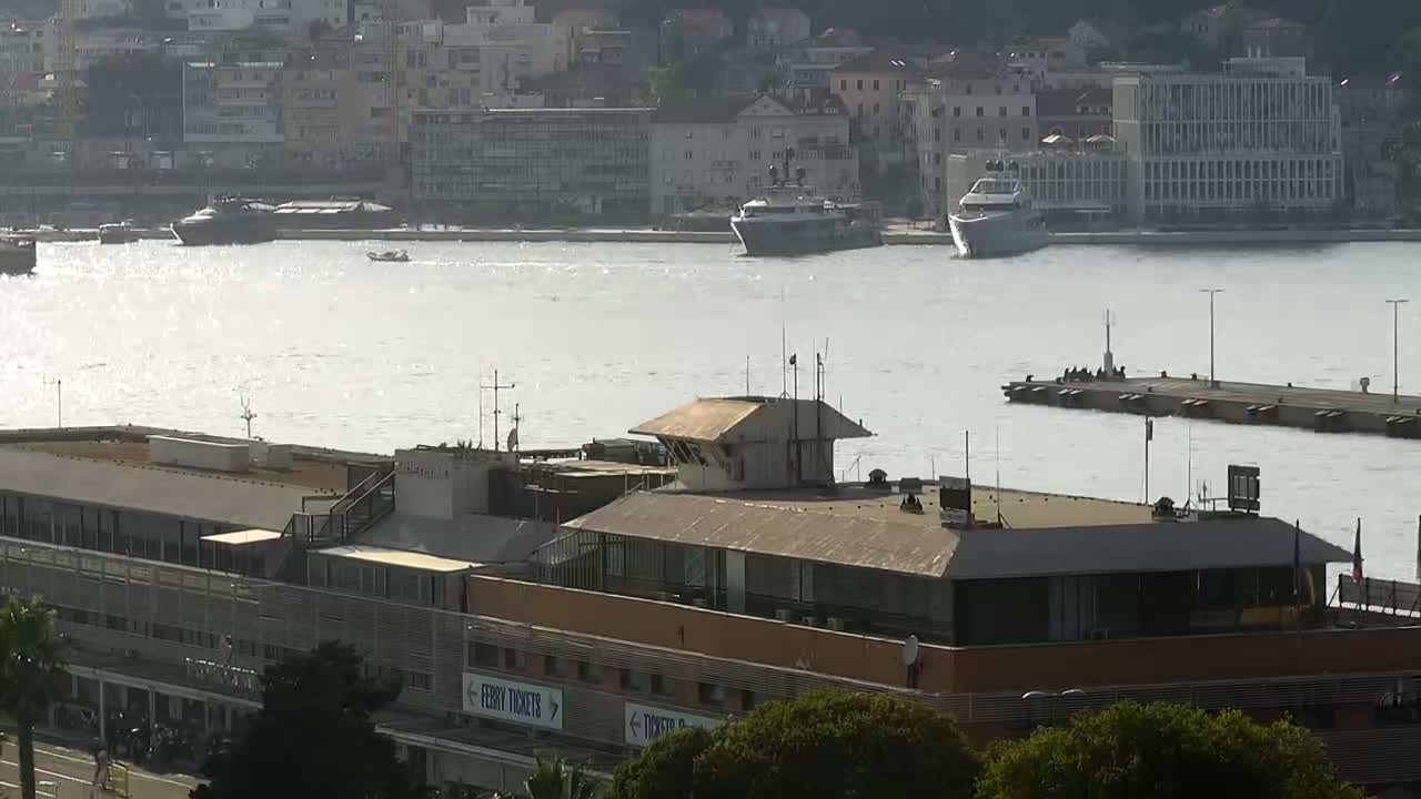 A harbor scene shows a ferry terminal building with a control tower, several boats in the water, and a city in the background on a somewhat hazy day.