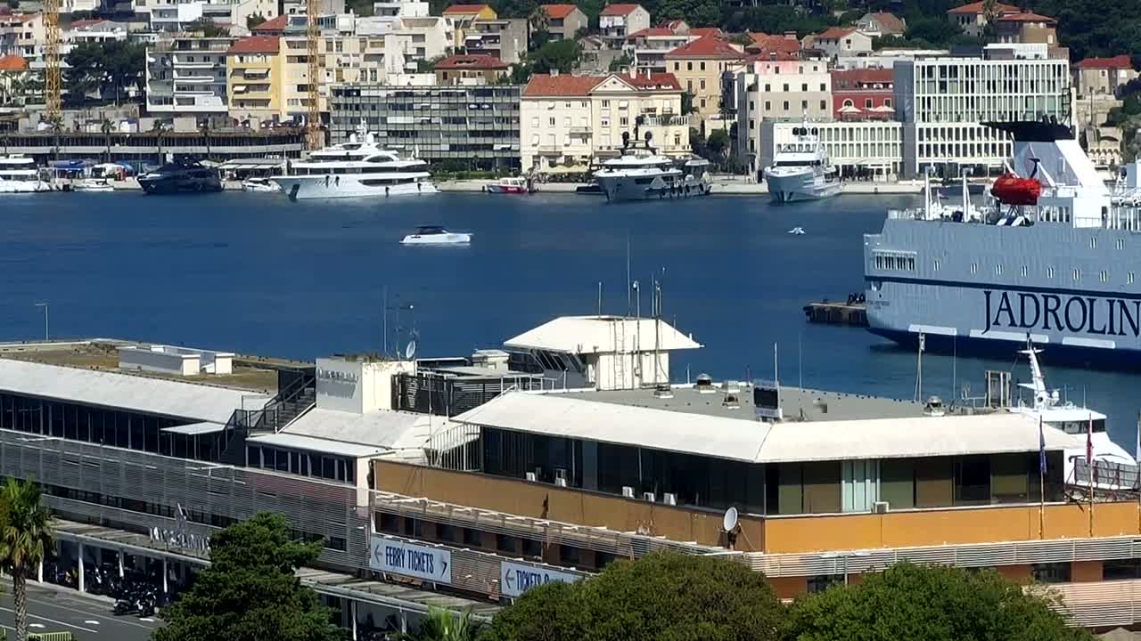 A ferry and several yachts are docked in a harbor next to a building, under sunny skies.