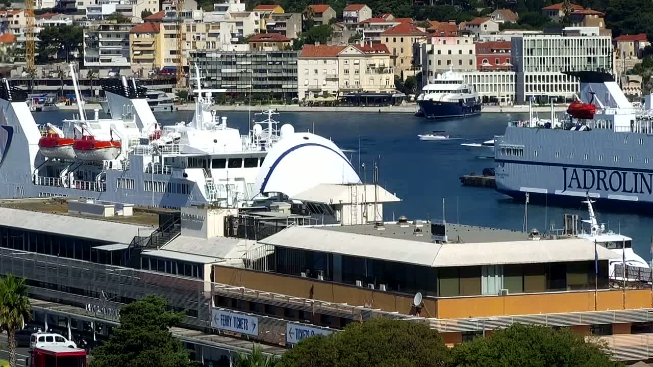A harbor scene on a sunny day shows several large passenger ships docked near a ferry terminal building, with a city in the background.