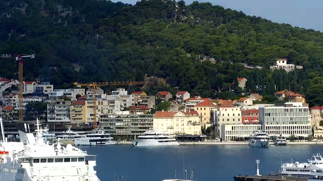 A waterfront town with numerous buildings is nestled at the base of a verdant hill on a clear, sunny day, with several boats and ships in the harbor.