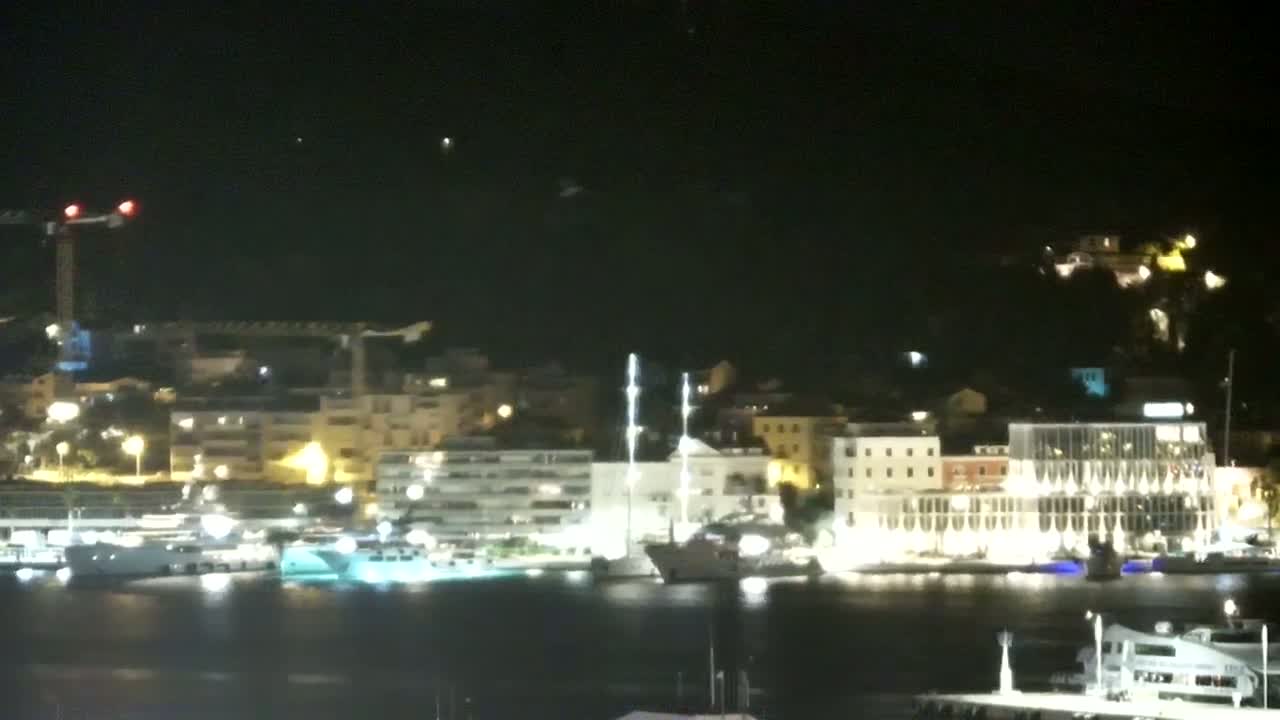 A waterfront town with numerous buildings is nestled at the base of a verdant hill on a clear, sunny day, with several boats and ships in the harbor.