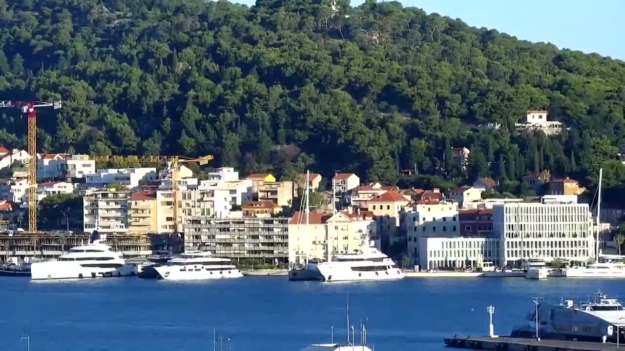 A waterfront town with numerous buildings is nestled at the base of a verdant hill on a clear, sunny day, with several boats and ships in the harbor.