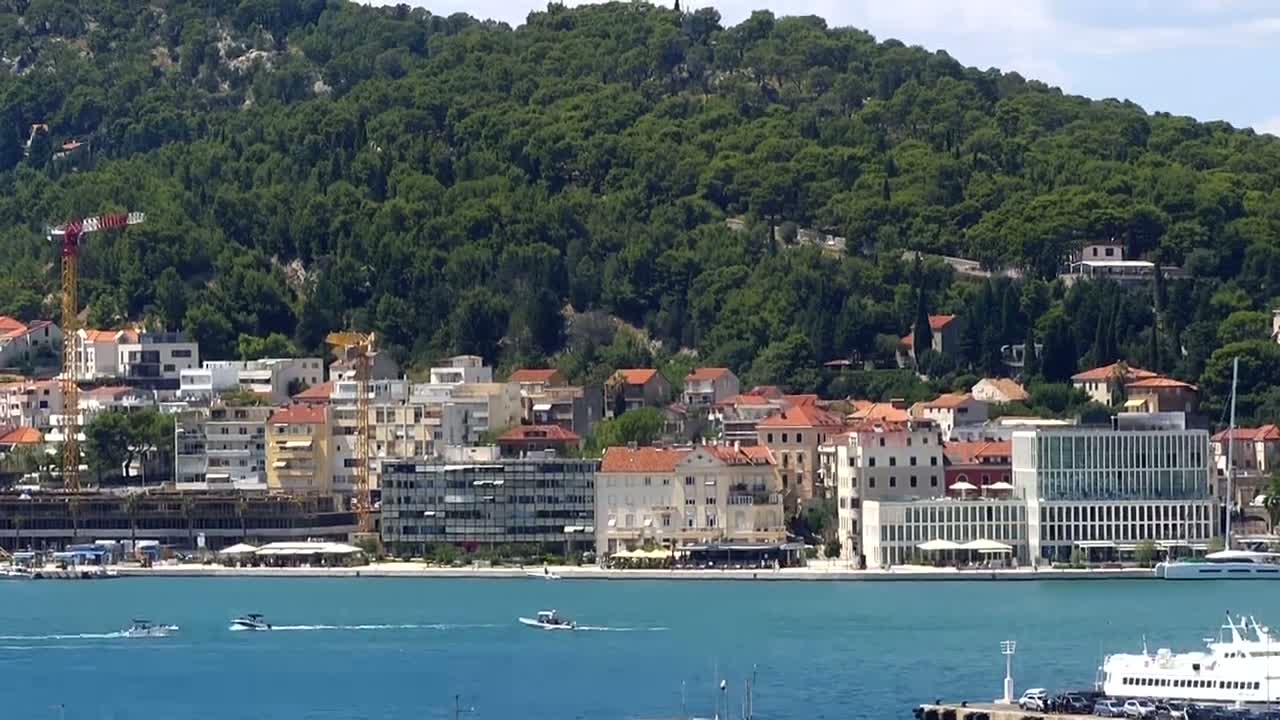 A waterfront town with numerous buildings is nestled at the base of a verdant hill on a clear, sunny day, with several boats and ships in the harbor.