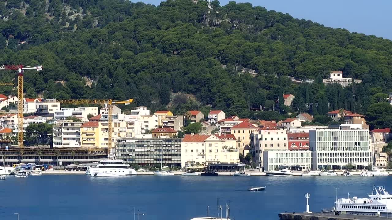 A coastal town with multi-story buildings and construction cranes sits at the base of a lush green hill on a sunny day.