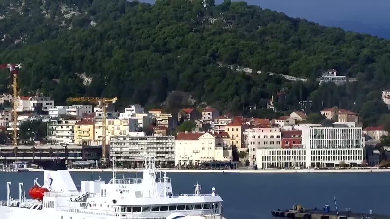 A large white ferry boat floats on blue water in front of a bustling coastal town with a mix of buildings and construction cranes, all set against a steep, densely forested mountain under a clear, sunny sky.