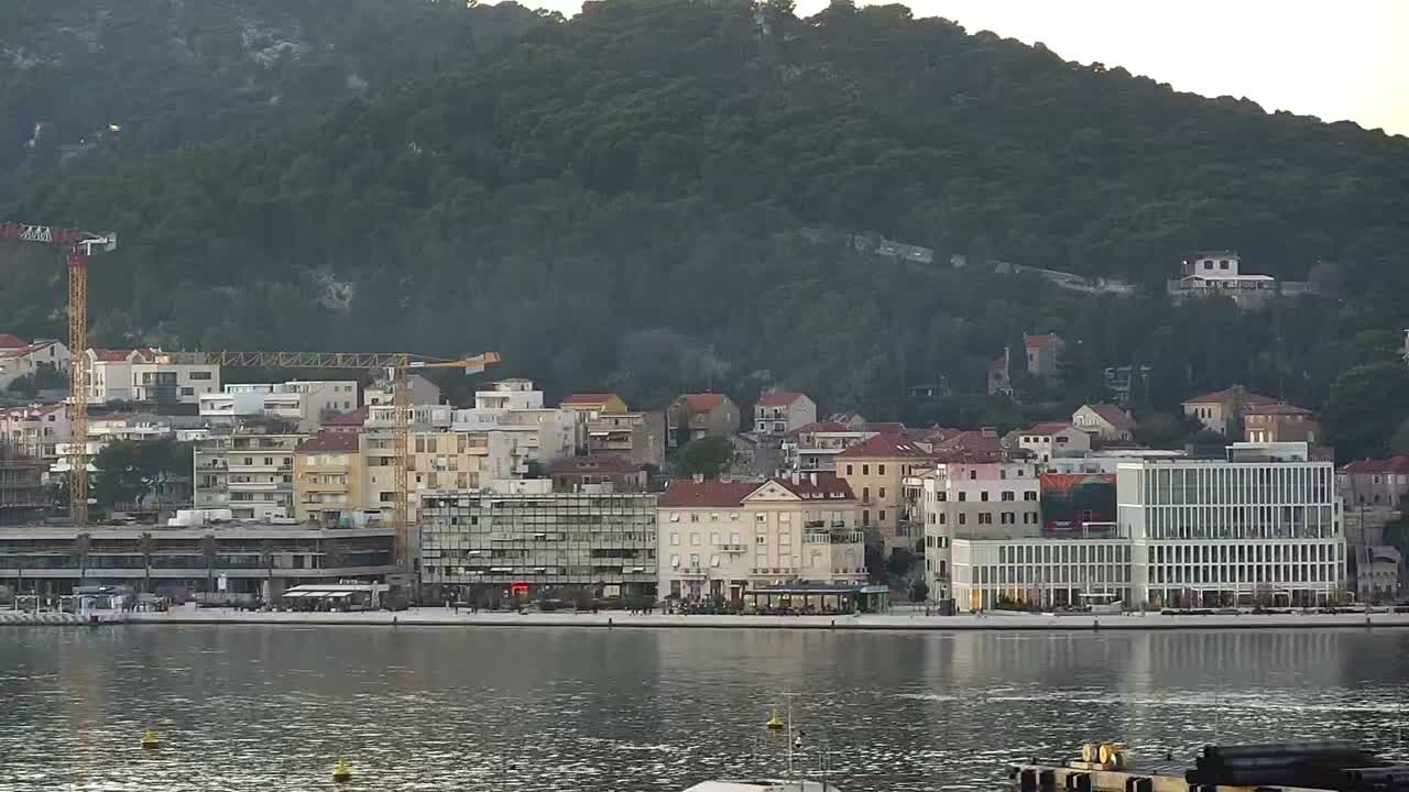 A large white ferry boat floats on blue water in front of a bustling coastal town with a mix of buildings and construction cranes, all set against a steep, densely forested mountain under a clear, sunny sky.