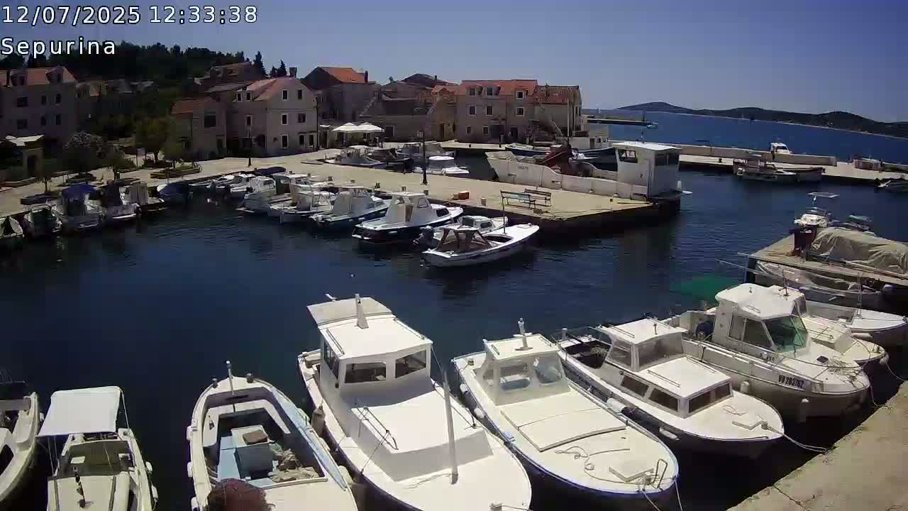 A sunny harbor filled with numerous boats is nestled beside a village of stone buildings on a clear day.