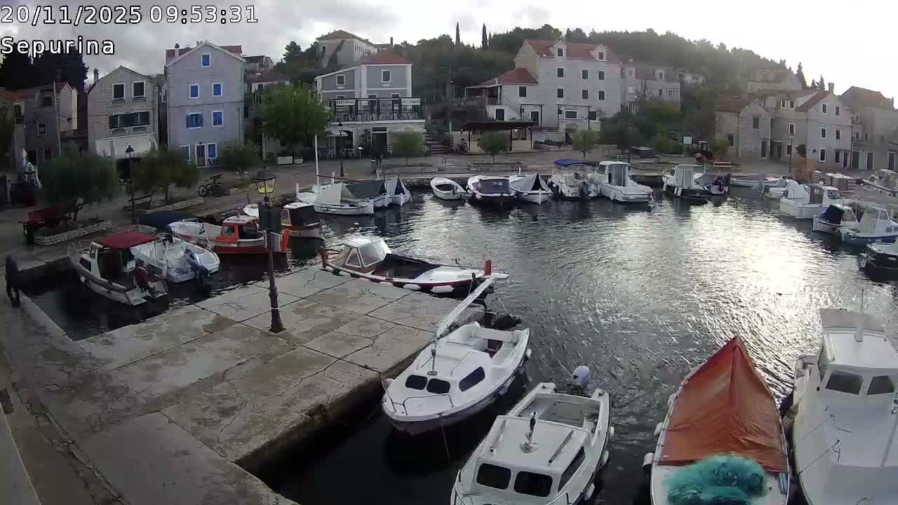 Numerous small boats are moored in a calm harbor framed by stone piers and multi-story houses climbing a hillside under an overcast sky.