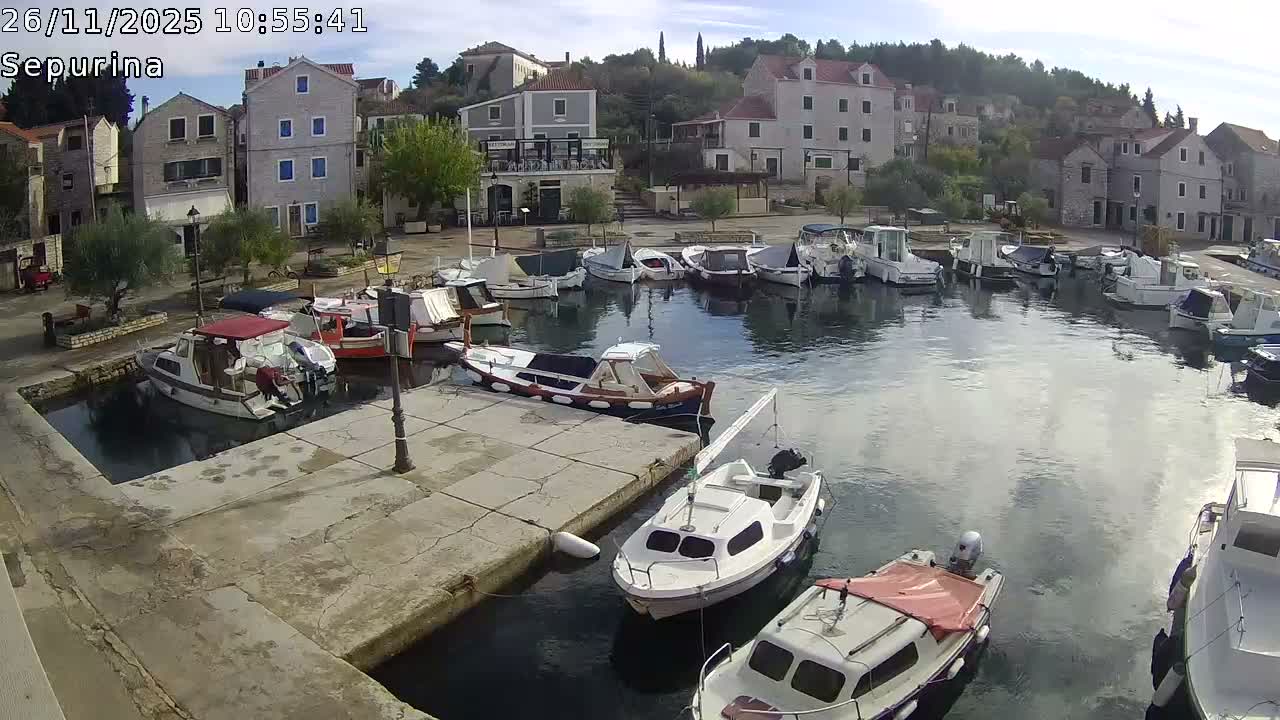 Numerous boats are docked in a calm harbor surrounded by historic buildings and green trees under a partly cloudy sky and fair weather.