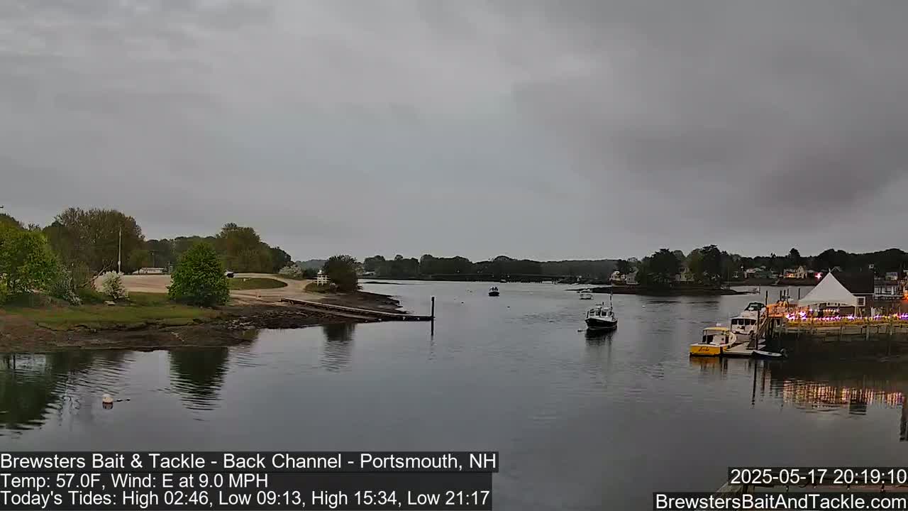 A calm river scene under an overcast sky shows several boats moored near a waterfront building with a tent and lights.