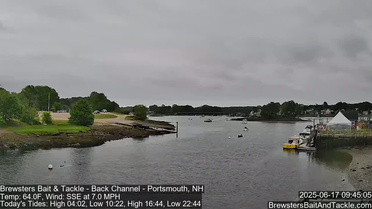 A calm, overcast day shows a waterway with several small boats, a shoreline with trees and buildings, and a white tent near a dock.