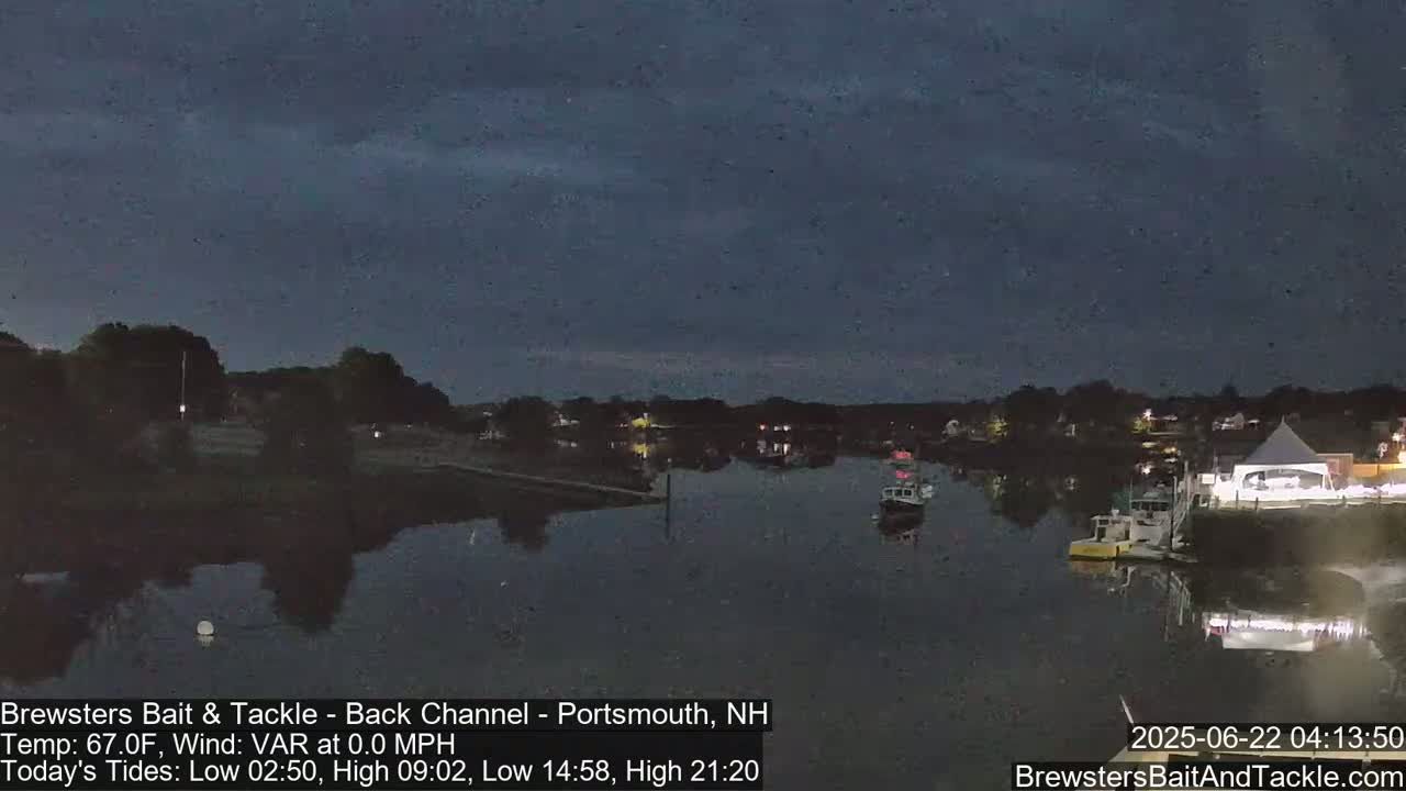 A calm, dark-blue evening scene shows a river with several boats moored near a shoreline with buildings and a large tent under a mostly clear sky.