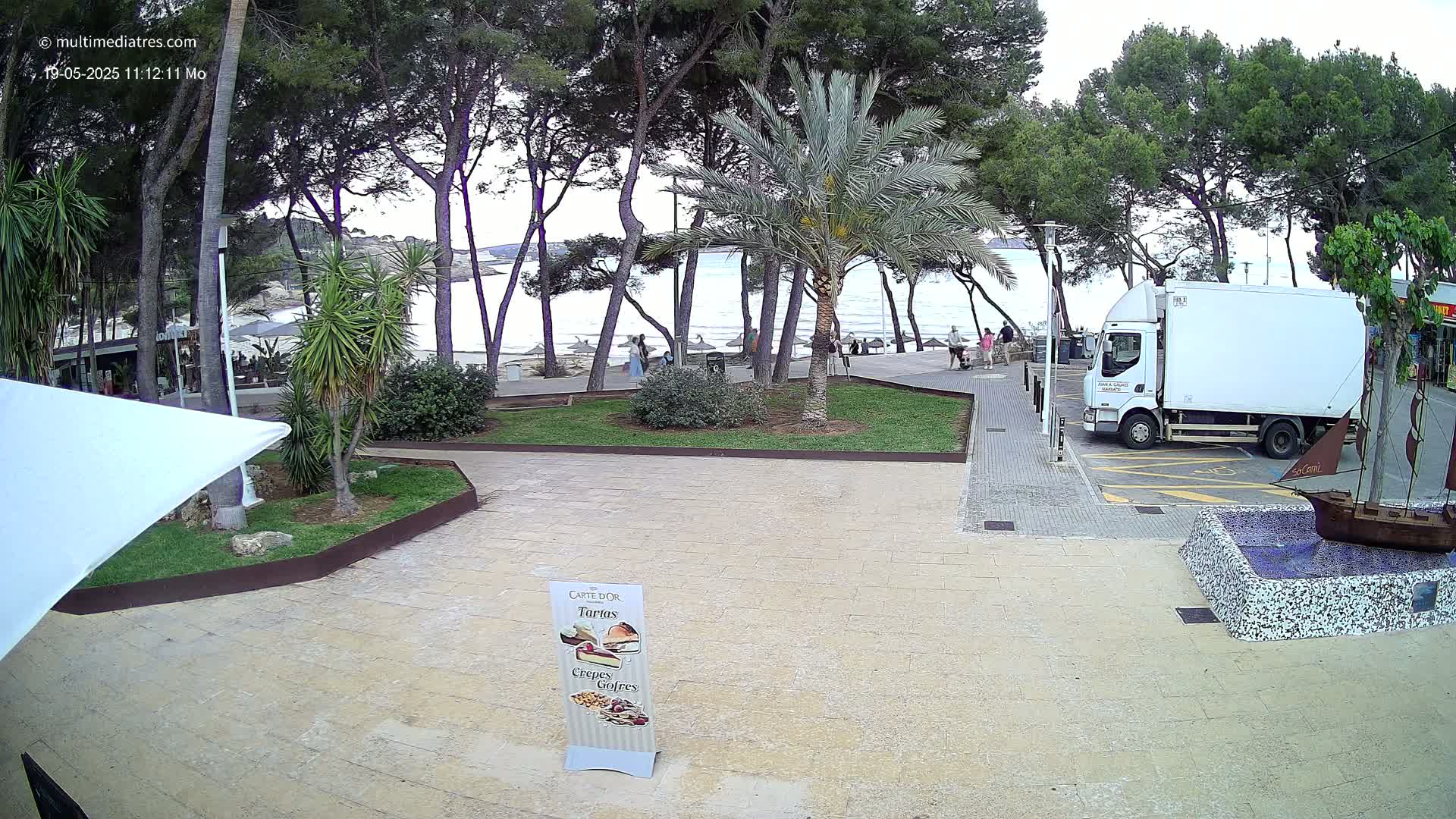 A paved area near a beach, partially shaded by trees and palm trees, with a white delivery truck parked nearby under an overcast sky.