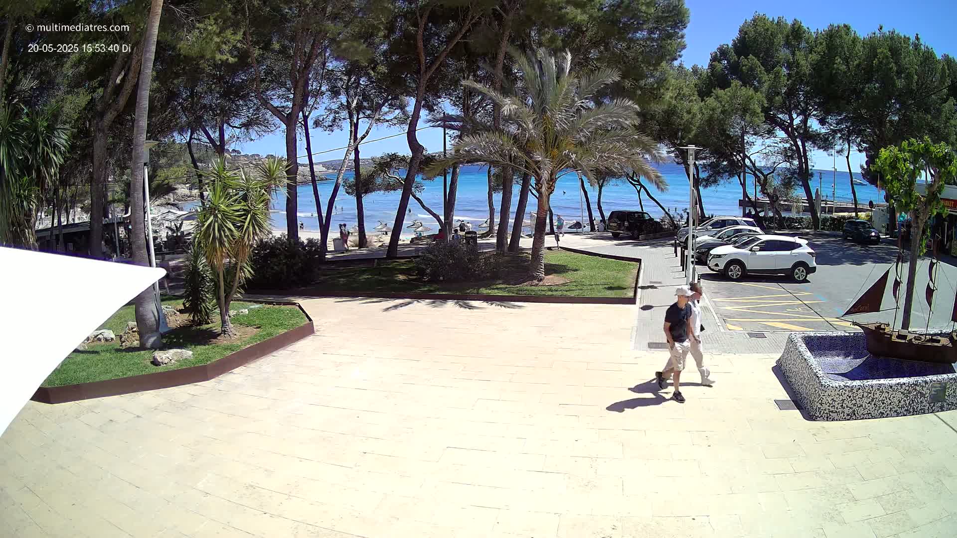 Two people walk on a paved area near a beach under a sunny sky, with cars parked in a lot and palm trees surrounding the scene.